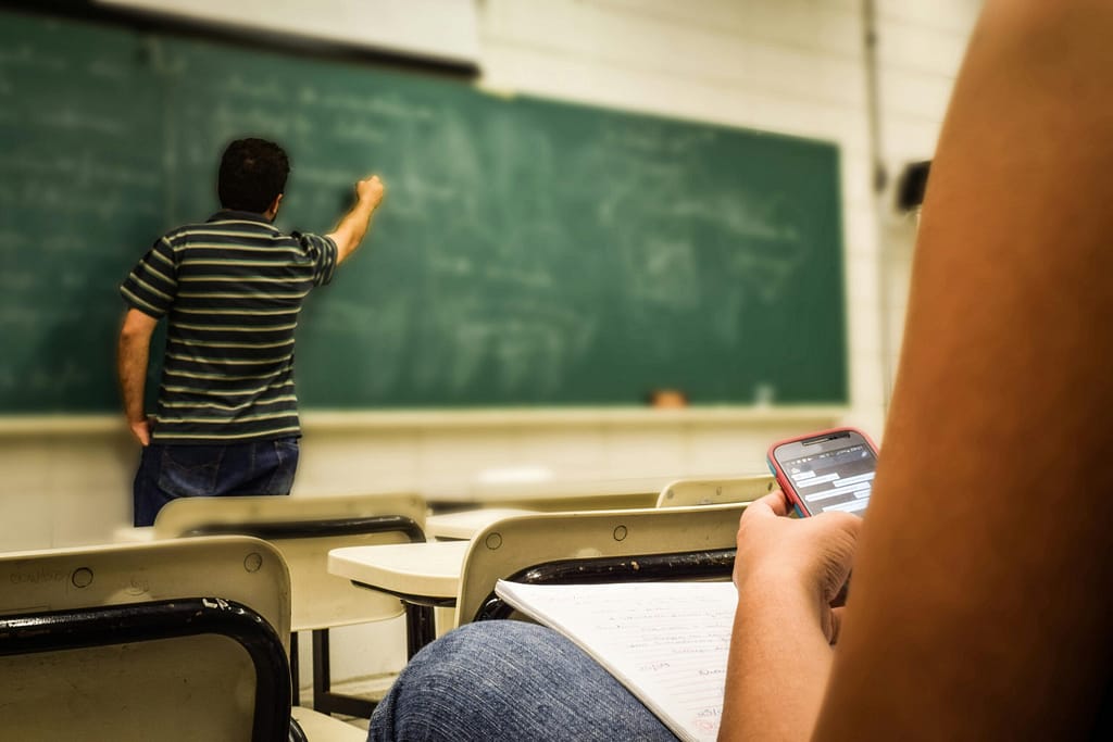 The Role of Technology in Education Today Student texting in a classroom while teacher is writing on the blackboard.